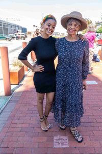 Two African-American women wearing dresses hug and smile while standing by a memorial brick.